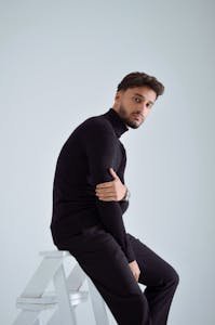 Stylish young man in black turtleneck posing on a stool with a minimalist background.